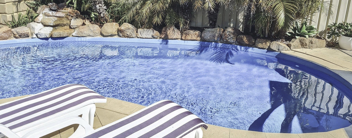 A swimming pool with blue water surrounded by tropical palm trees, rocks, and two white lounge chairs with striped cushions